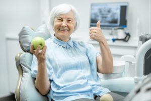 senior woman in dental chair giving thumbs up and holding green apple 